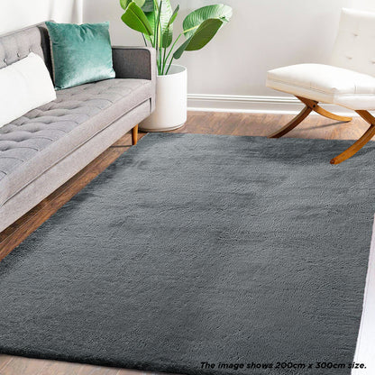 Azaliya Dark Grey Faux Rabbit Fur Rug 200x300cm lies in a modern living room, bordered by a tufted light-gray sofa and a white armless chair, enhancing the cozy, minimalist aesthetic.