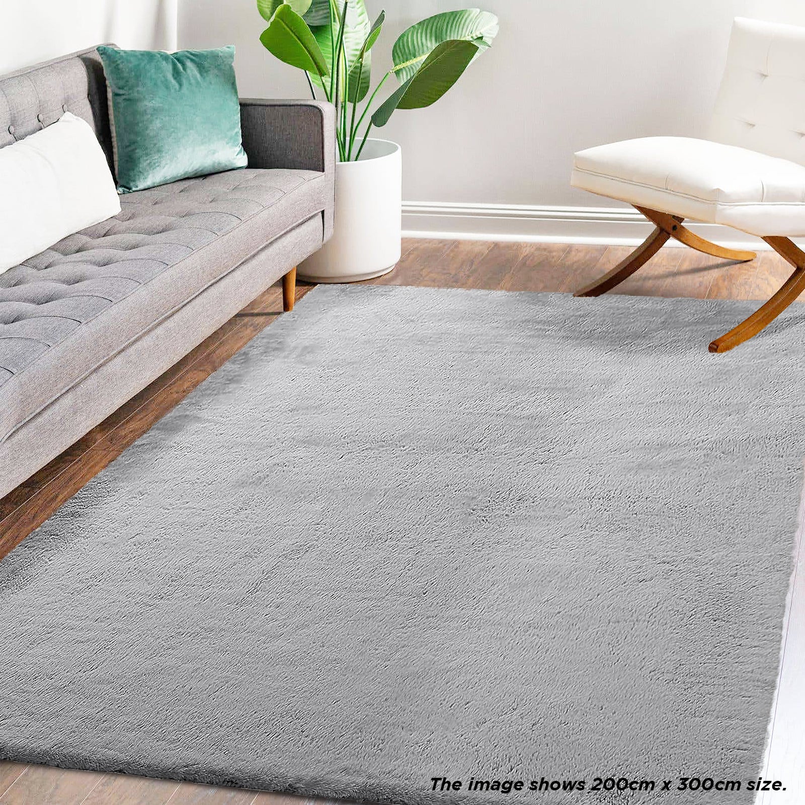 Azaliya 200x300cm Faux Rabbit Fur Floor Rug in Silver, styled in a modern living room with a gray mid-century sofa, green pillow, white planter, and wooden-legged lounge chair.