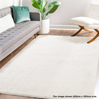 Azaliya 200x300cm Faux Rabbit Fur Floor Rug in Cream White in a modern living room with a gray tufted sofa, potted plant, and white armless chair on warm wood flooring.