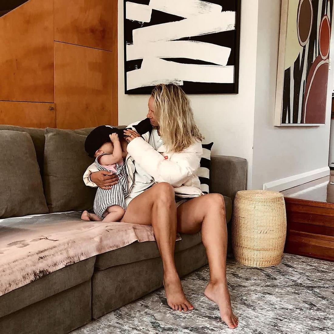 A woman sitting on a couch holds a baby, with the Foldvik Art Contemporary Rug 160x230cm visible beneath them, featuring a light tribal-inspired pattern in greens and creams, enhancing the modern decor.