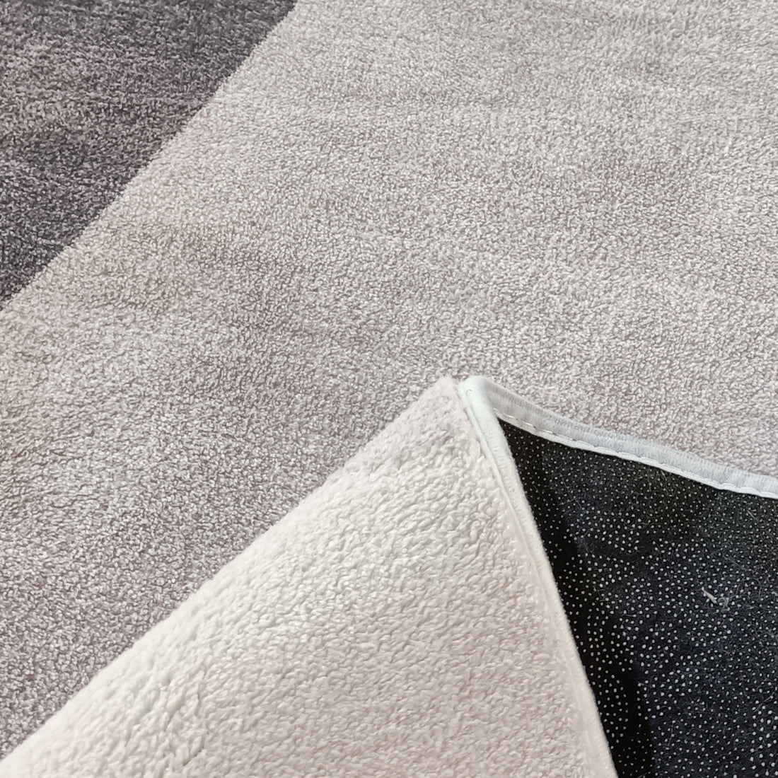 Glote 120x180cm Small Abstract Indoor Floor Rug on grey carpet, corner flipped to show black non-skid backing with white rubberized dots, showcasing its textured surface and subtle abstract leaf pattern.