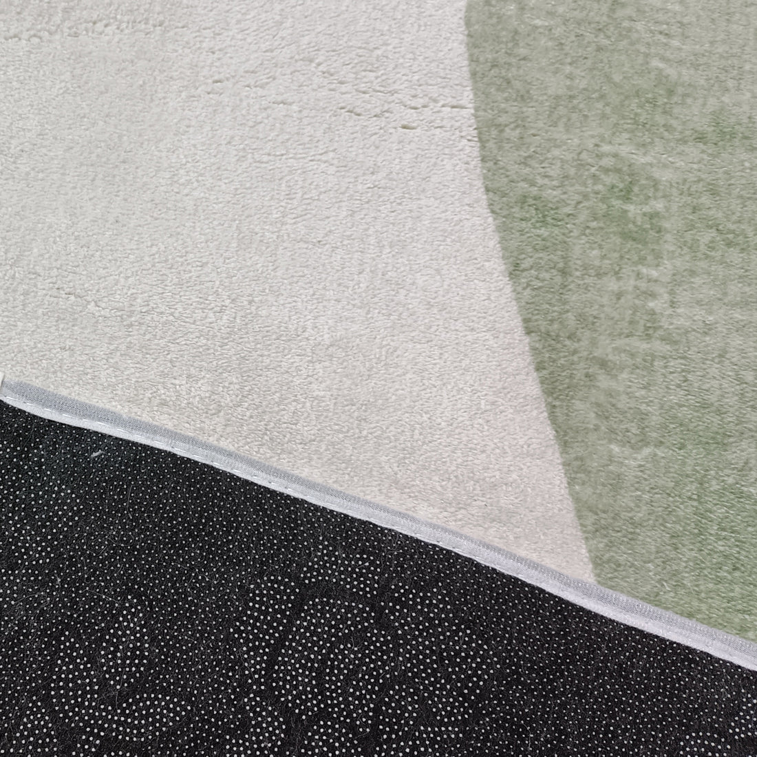 Close-up of Gonas 120x180cm Small Abstract Indoor Floor Rug showing off-white and pale green sections with sewn-on binding, and black dotted non-slip backing peeking beneath.