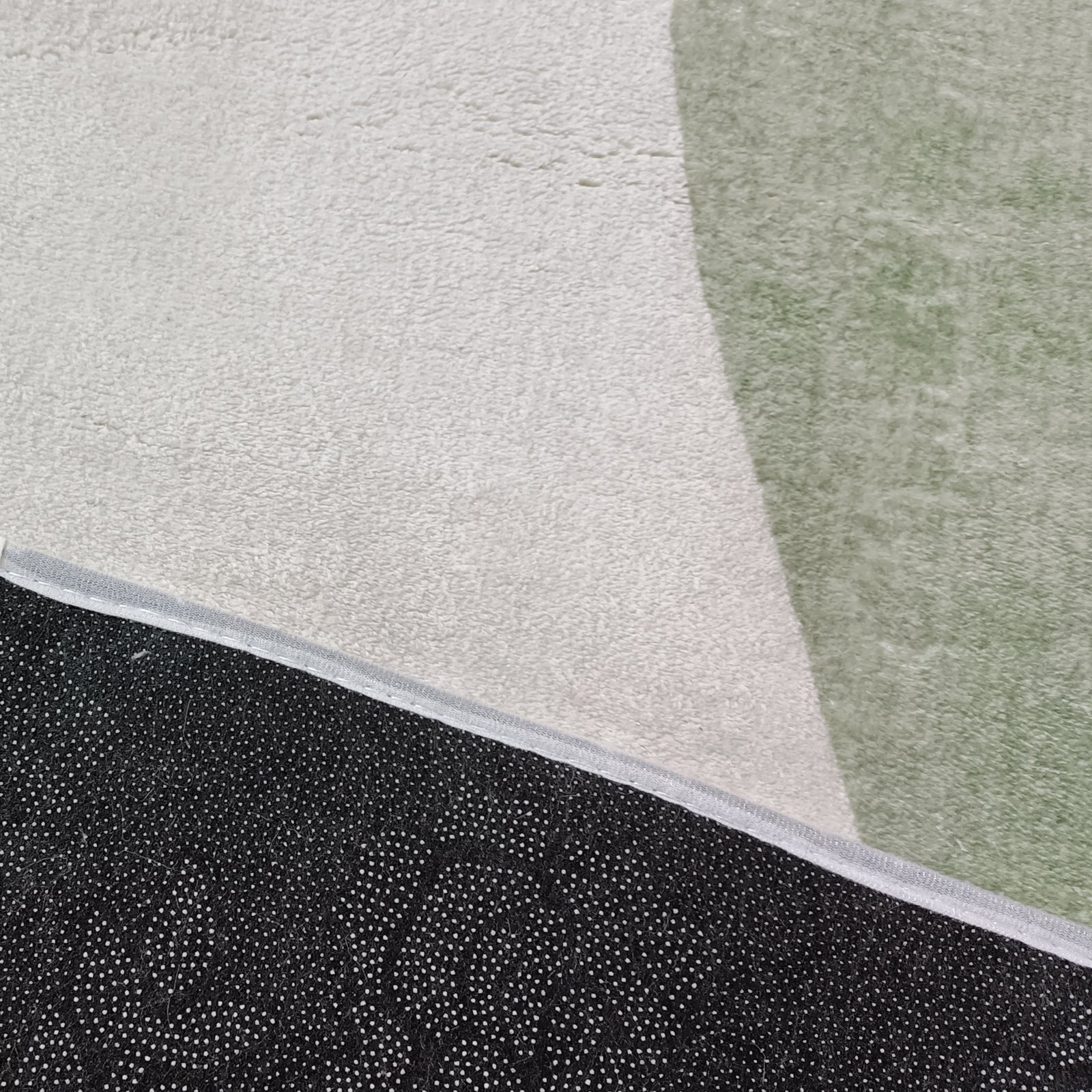 Close-up of the Gonas 160x230cm Medium Abstract Indoor Floor Rug, showcasing an off-white surface with a pale green shape and a black, dot-patterned non-slip backing for stability.
