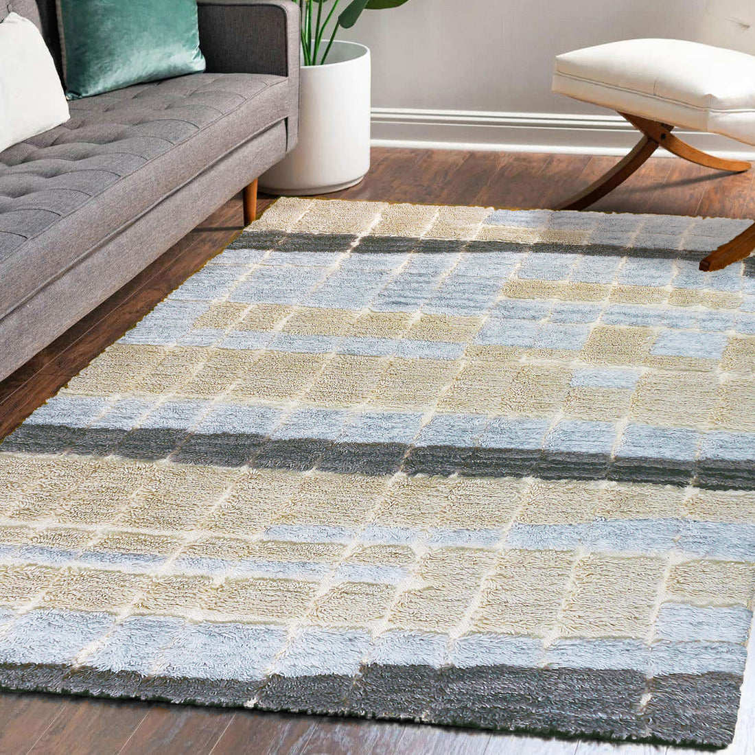Isaac 160x230cm Abstract Wool Blend Handwoven Shag Rug displayed on dark hardwood floor, featuring a checker-style pattern with muted tones, complementing a mid-century-inspired gray sofa and adjacent white planter.