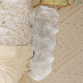 Kalina 60x180cm Shaggy Fluffy Fur Rug draped on light-toned herringbone wooden floor beside a bed with floral linens, adding a plush, cozy texture to the bedroom setting.