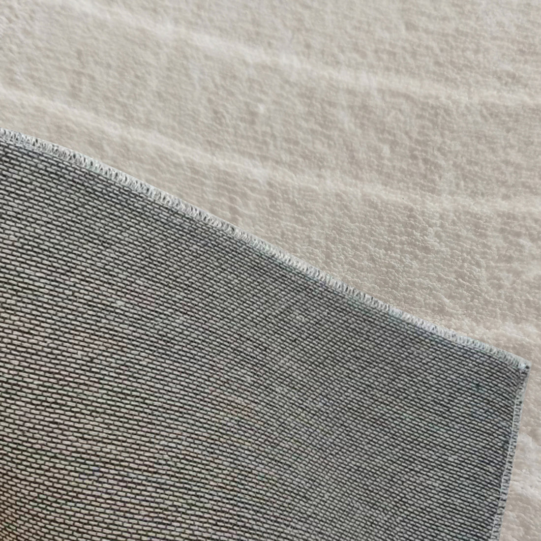 Close-up of Lita 120x180cm Polyester Tufted Modern Abstract Rug corner, showcasing its grey woven texture atop a soft off-white surface, highlighting its intricate stitching and modern design.