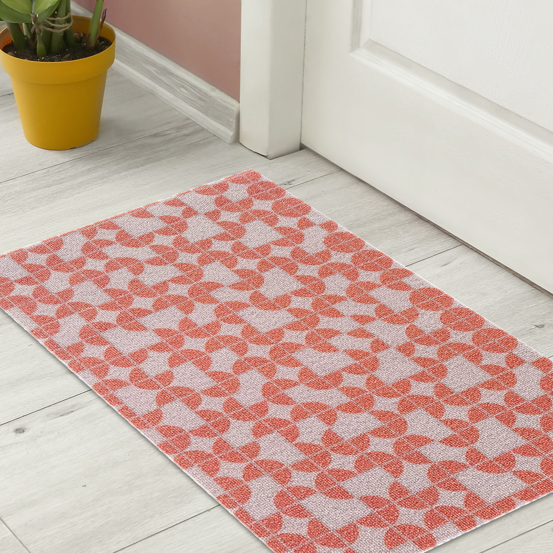 Macy 45x75cm Modern Outdoor Doormat in Light Terracotta with a geometric orange pattern, leaning against a white door beside a mustard-yellow pot with a leafy plant.
