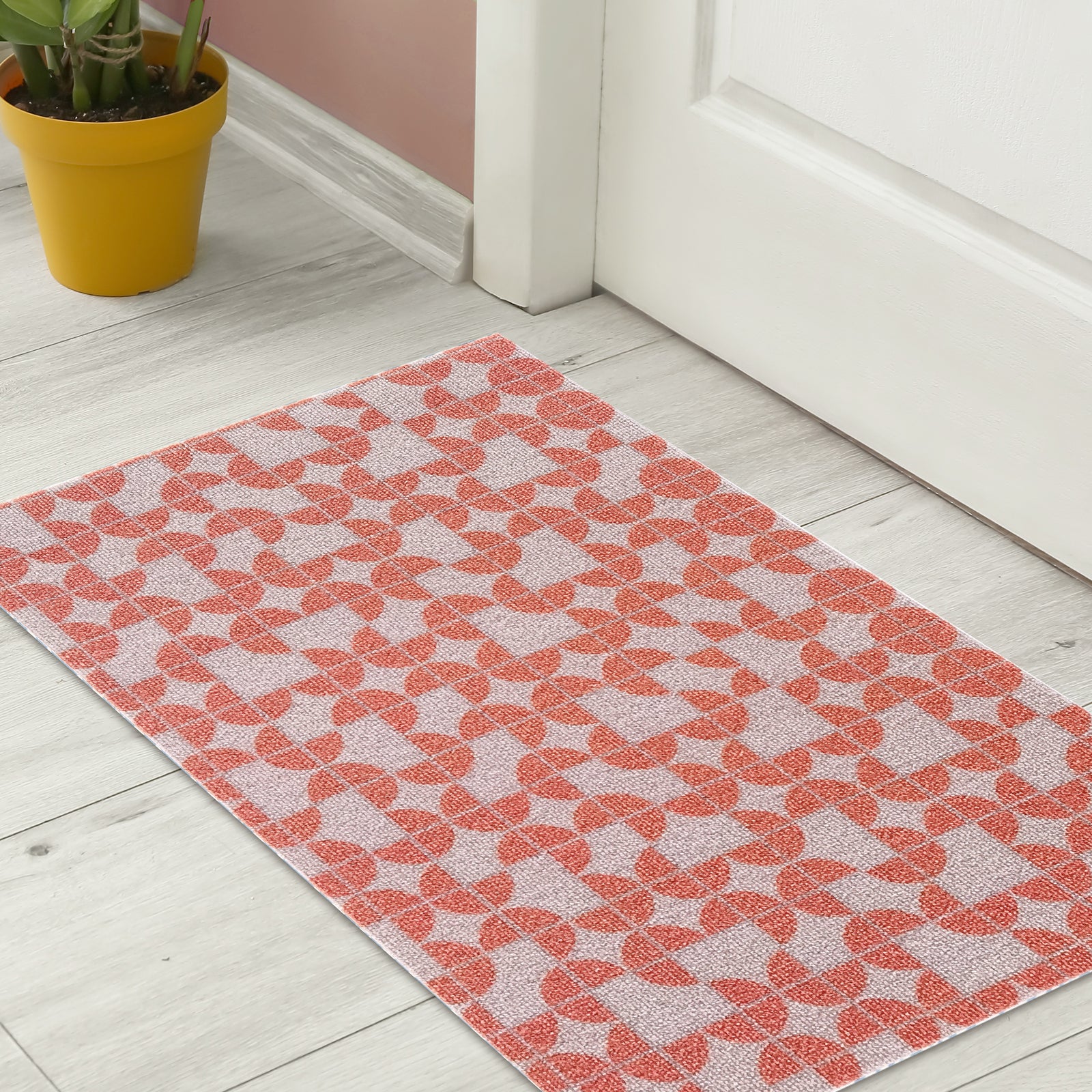 Macy 45x75cm Modern Outdoor Doormat in Light Terracotta with a geometric orange pattern, leaning against a white door beside a mustard-yellow pot with a leafy plant.