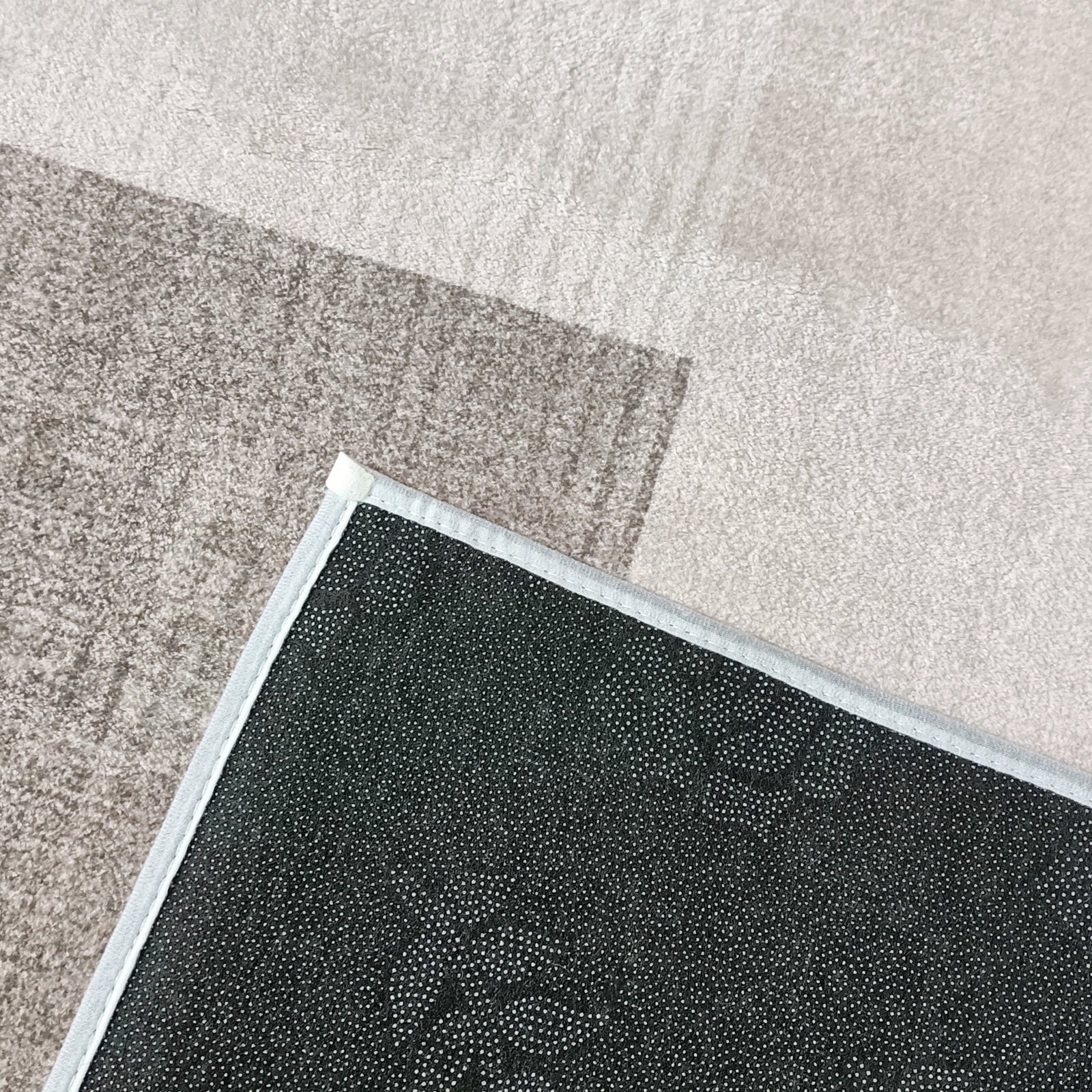 Close-up of the Orsa 120x180cm Small Contemporary Indoor Floor Rug, showing its flipped corner with a black, dot-textured, non-slip underside on a beige carpet.