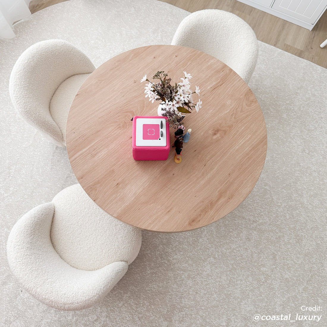 Overhead view of Pavla 200 x 300cm Oval Soft Rug in Cream White under a light-oak table, surrounded by three boucle chairs, featuring a white vase, pink Toniebox, and figurines on top.
