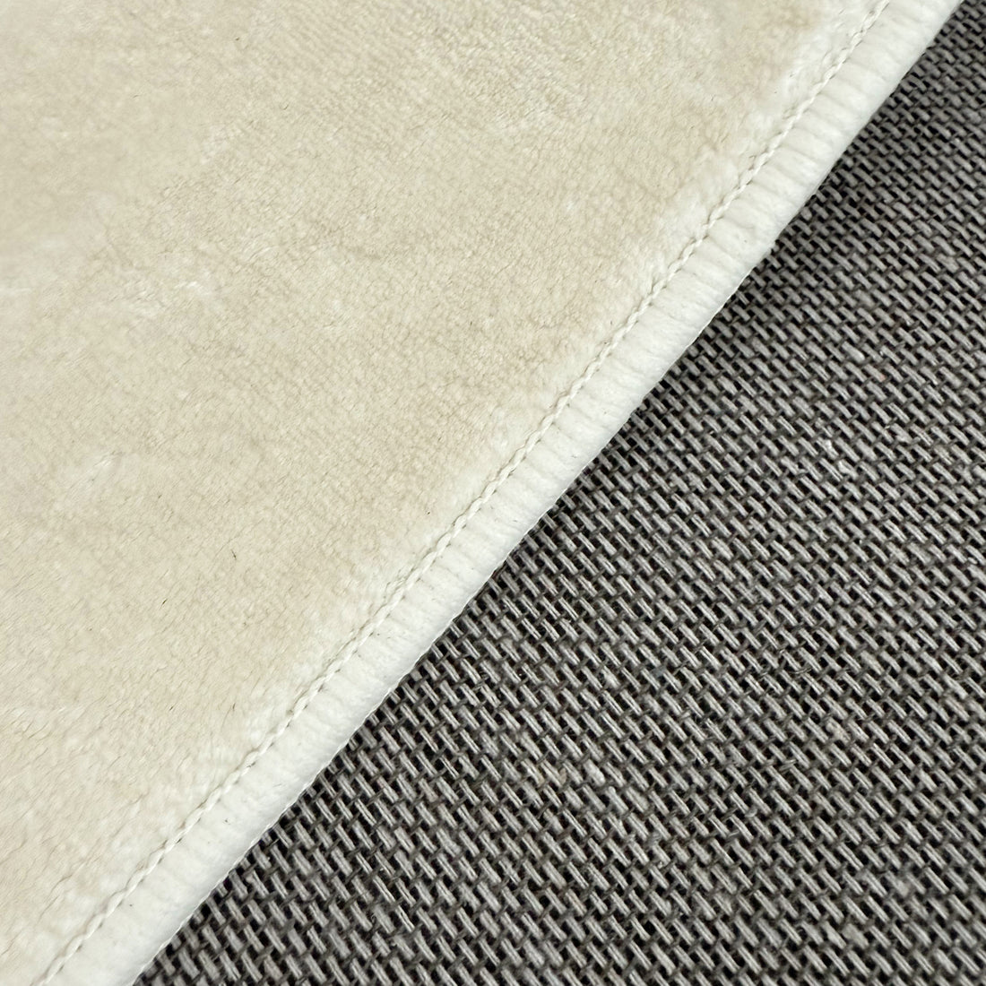 Close-up of Pavla Oval Shaggy Rug - Beige, showcasing its plush, cream fabric with stitched edge alongside a contrasting gray, coarse, basket-weave texture, highlighting the rug's luxurious, soft texture.