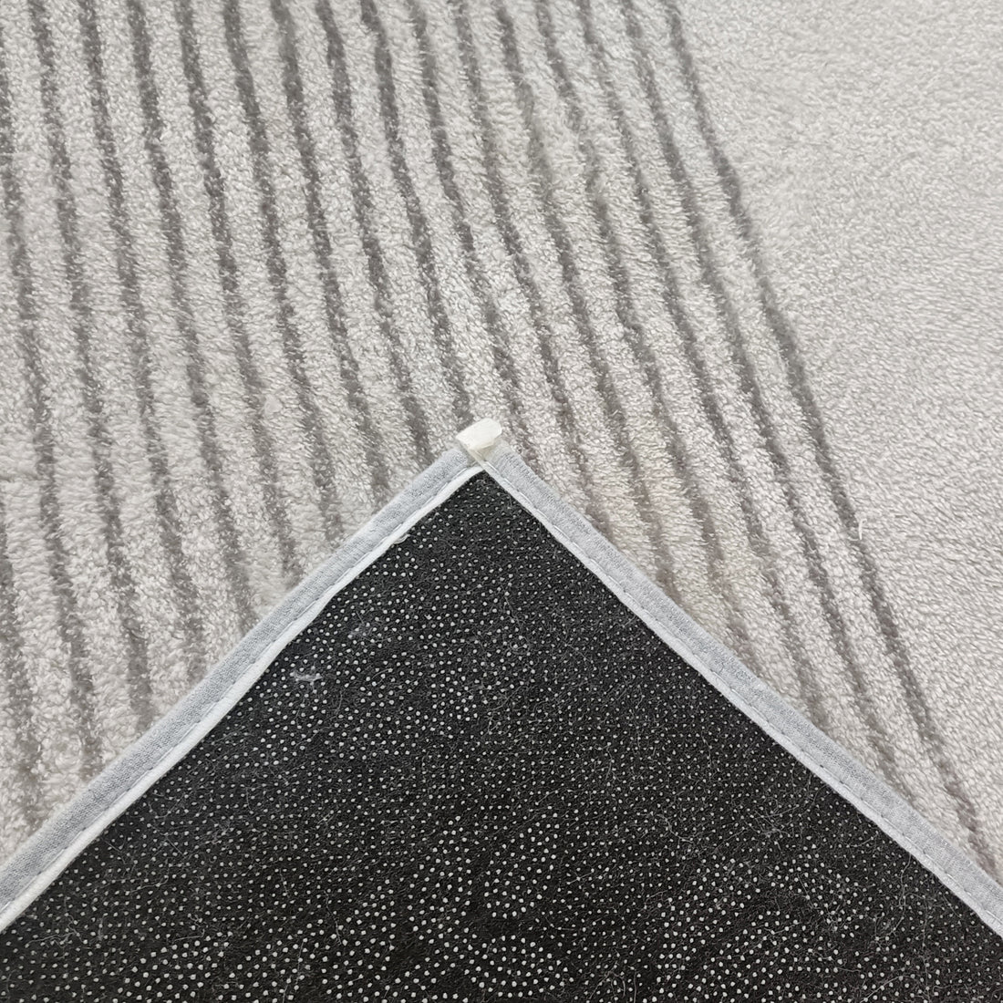 Ramvik 160x230cm Abstract Indoor Floor Rug with light gray, subtly striped surface, reveals black, dotted non-slip backing, displayed on a carpeted floor, highlighting its anti-slip functionality and modern design.