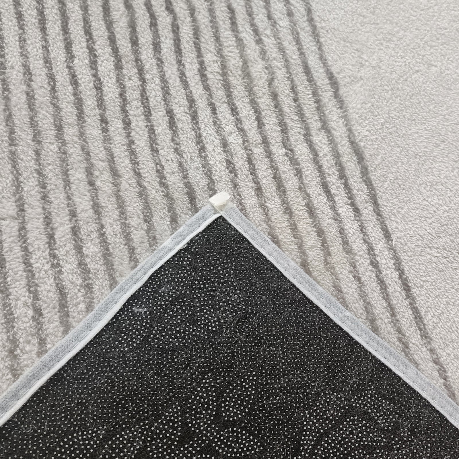 Ramvik 160x230cm Abstract Indoor Floor Rug with light gray, subtly striped surface, reveals black, dotted non-slip backing, displayed on a carpeted floor, highlighting its anti-slip functionality and modern design.