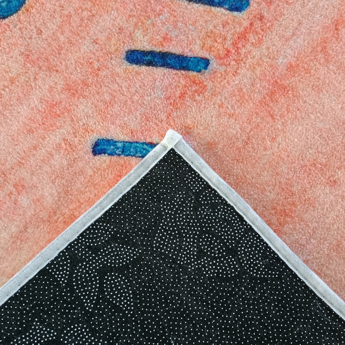 Close-up of the Rokke 160x230cm Kids Modern Abstract Indoor Floor Rug showing its fuzzy peach-and-blue pattern with white edge binding, and a lifted corner revealing black, anti-slip backing with grip dots.
