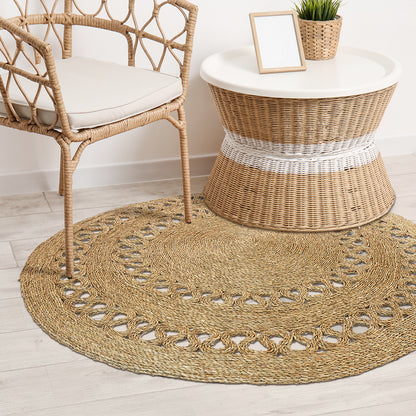 Sandra Brown Seagrass Rug 180cm, featuring intricate braided design and decorative looped edging, complements a minimalist boho interior with a rattan armchair and wicker side table arrangement.