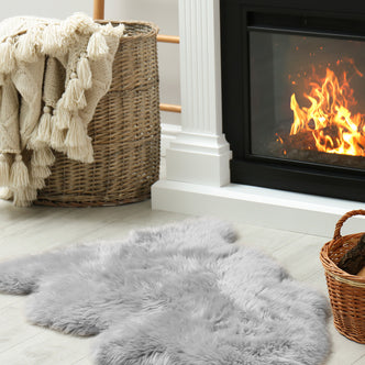 Silvanus 60x90cm Shaggy Soft Fluffy Fur Rug in Grey lies on a pale wood floor beside a wicker basket with a knit throw, near a small basket of firewood by the fireplace.