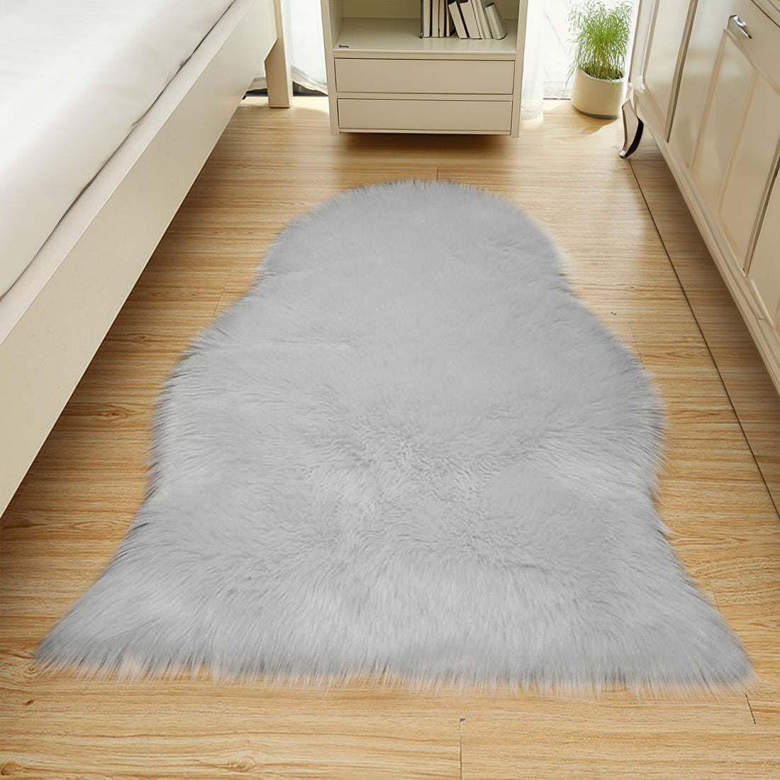 Silvanus 60x90cm Shaggy Soft Fluffy Fur Rug in Grey, plush and faux-fur, lies on light-wood flooring, beside a white bed frame and cream dresser, enhancing the cozy bedroom corner.