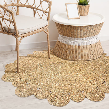 Una 180cm Seagrass Rug - Brown displayed under a boho-inspired setting with a woven rattan armchair, drum-shaped side table, and a cozy, braided design featuring scalloped edges.