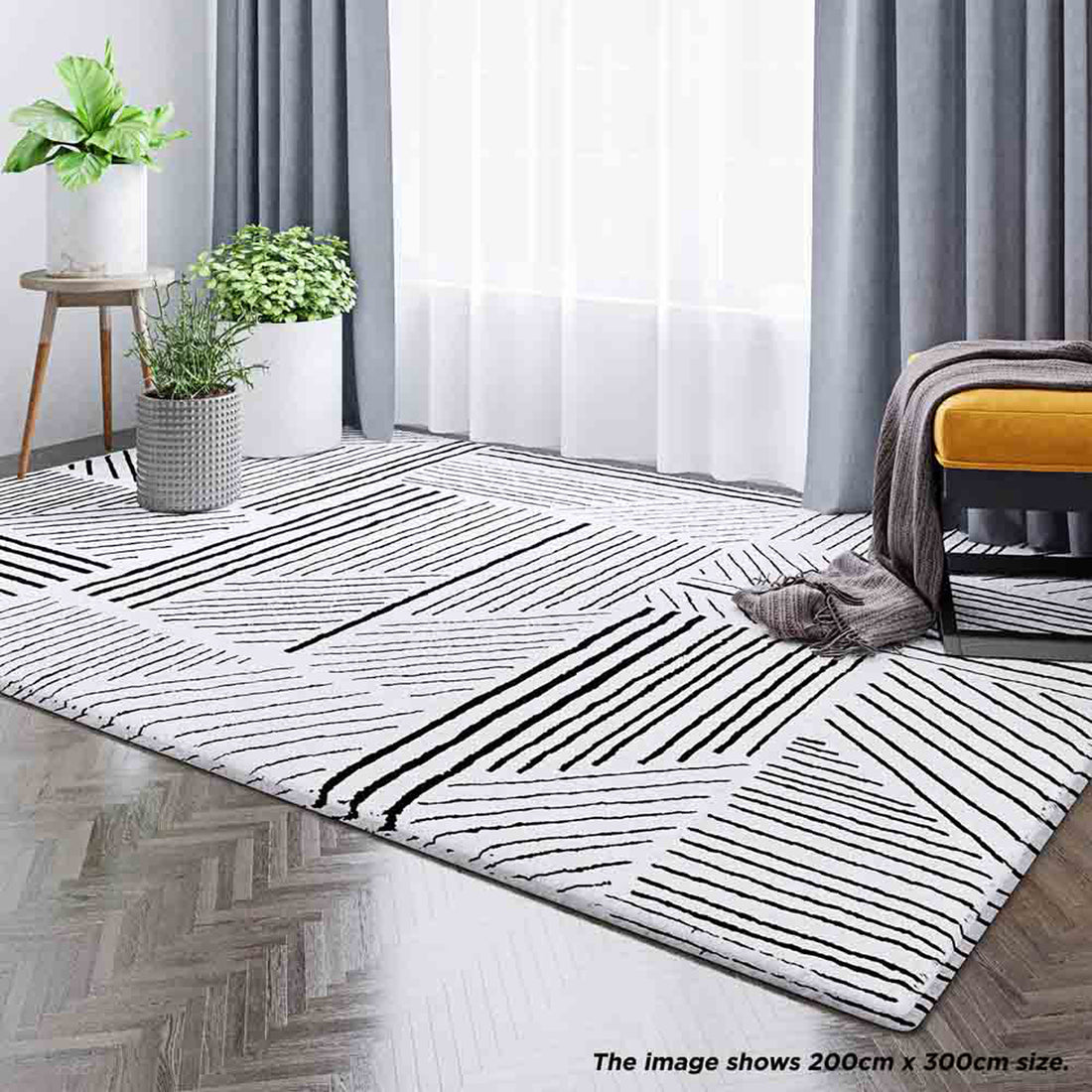 Vikki 200x300cm Abstract Contemporary Floor Rug with black-and-white geometric pattern, enhancing a modern room with potted plants, sheer curtains, and a mustard-yellow ottoman on herringbone wood flooring.