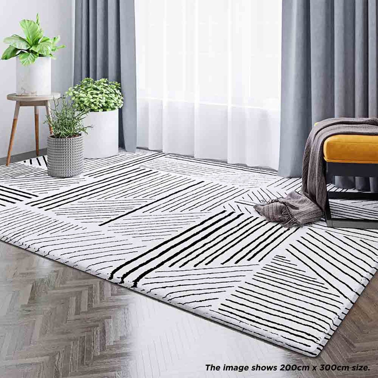 Vikki 200x300cm Abstract Contemporary Floor Rug with black-and-white geometric pattern, enhancing a modern room with potted plants, sheer curtains, and a mustard-yellow ottoman on herringbone wood flooring.