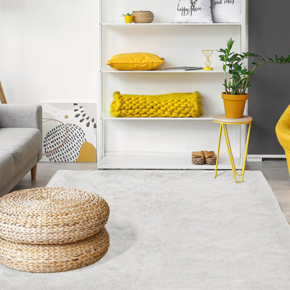 The Zubuin 180x300cm Shaggy Plush Rectangular Rug in White anchors a cozy living room with its soft, luxurious texture, complementing modern decor elements like a round woven pouf and shelving unit.