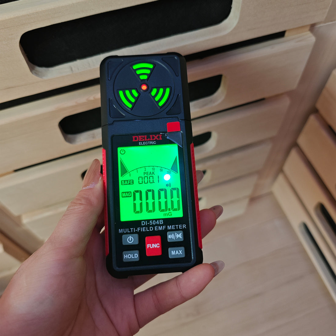 Hand holding a DELIXI DI-504B EMF meter displaying 000.0 mG reading in front of wooden sauna slats