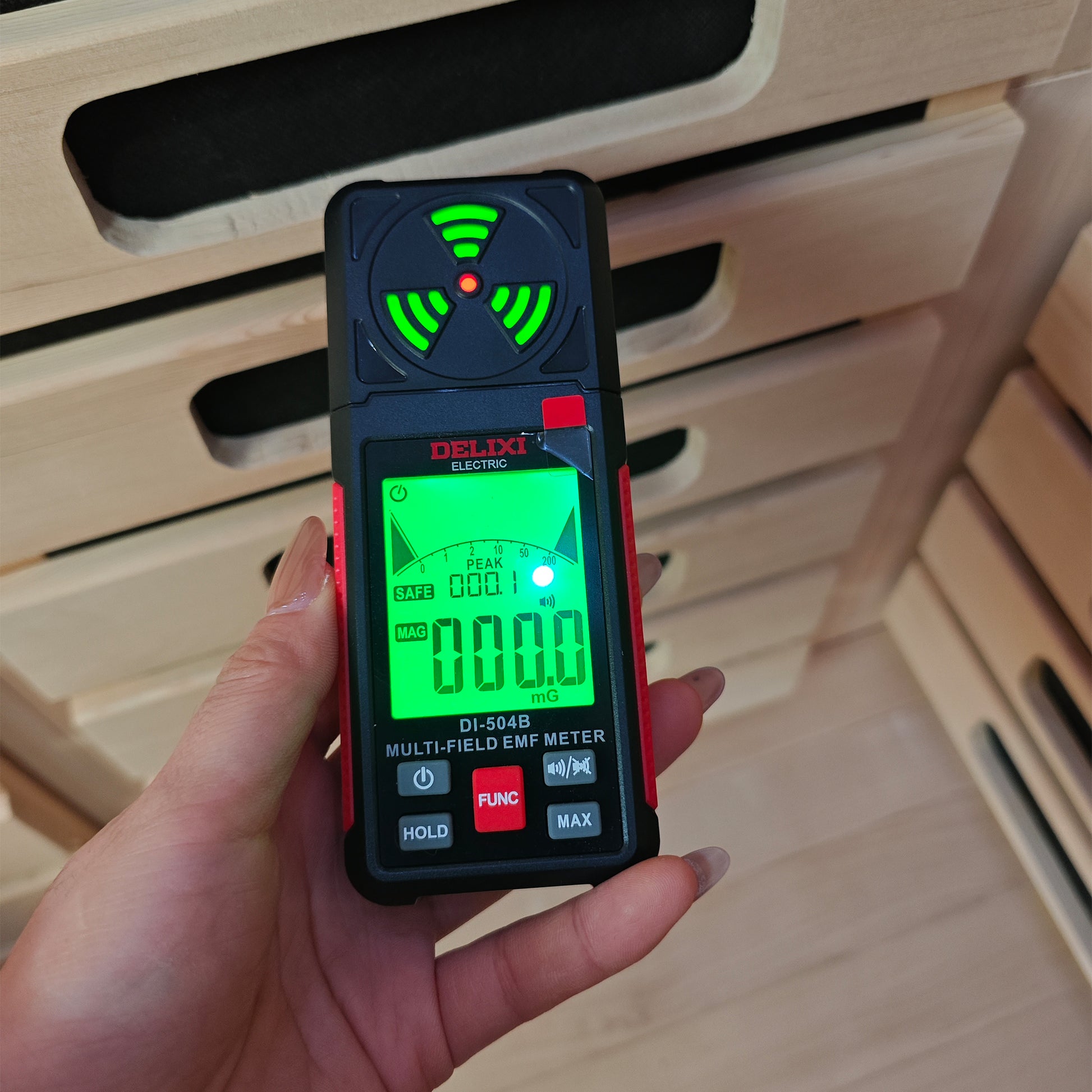 Hand holding a DELIXI DI-504B EMF meter displaying 000.0 mG reading in front of wooden sauna slats