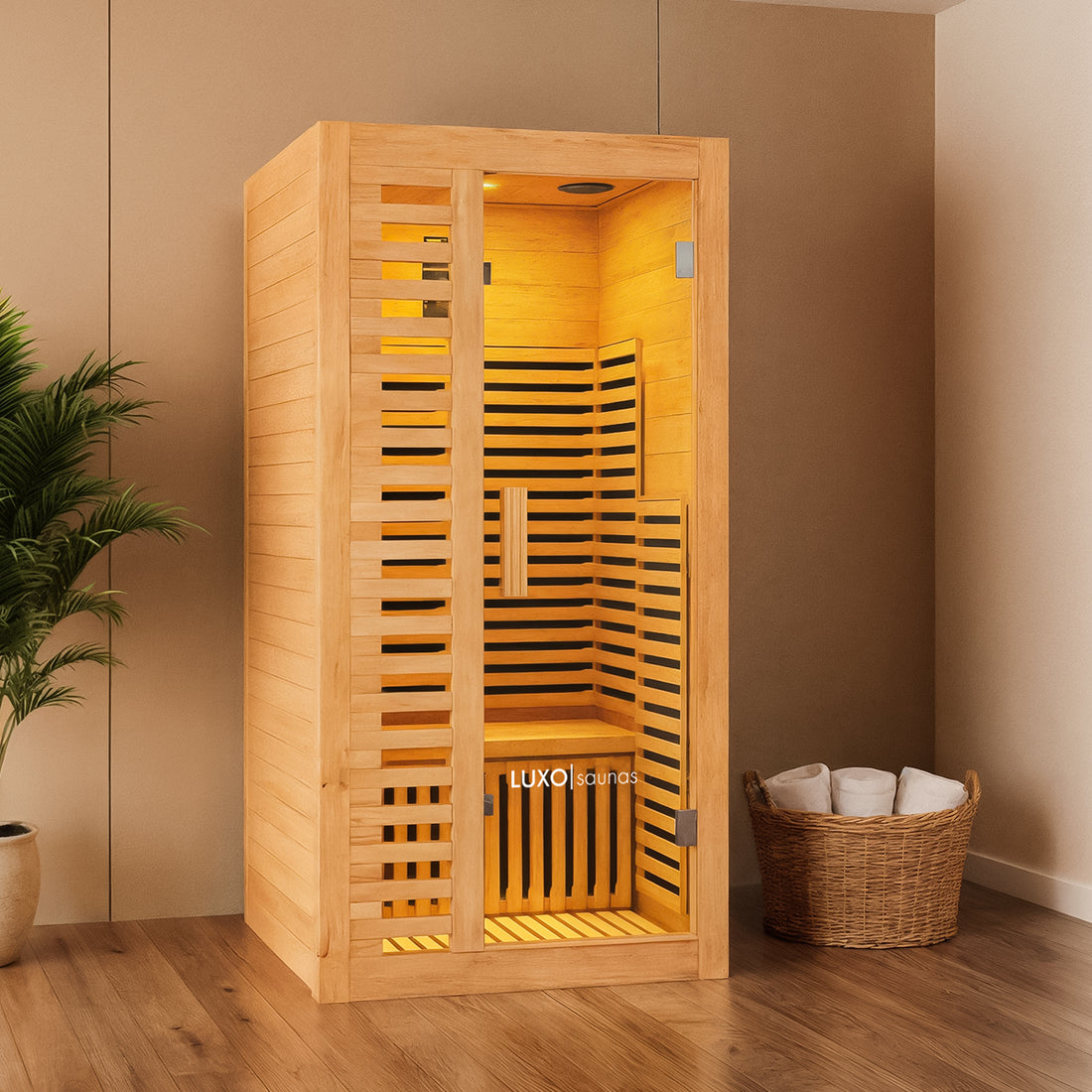 Harlow 1 Person Carbon Fibre Infrared Personal Indoor Sauna with light-colored timber slats, glass front door, and interior bench seating, softly illuminated, with a nearby potted plant and basket of towels.