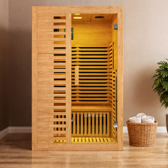 Harlow 2 Person Carbon Fibre Infrared Personal Indoor Sauna, featuring a wooden cabin with a clear door, single bench, and a warm glow, complemented by a wicker basket with towels and a potted plant nearby.