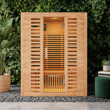 Harlow 3 Person Carbon Fibre Infrared Personal Indoor Sauna with horizontal cedar slats and full-height glass door, set on river stones, surrounded by a green wall, tropical plant, and ottoman.