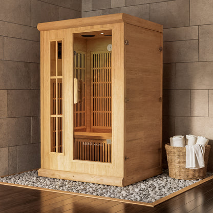 Hirsi 2 Person Carbon Fibre Infrared Indoor Home Sauna, crafted from light-wood, set on white stones, featuring vertical slatted panels, a bench, and a control panel, with towels in a wicker basket nearby.
