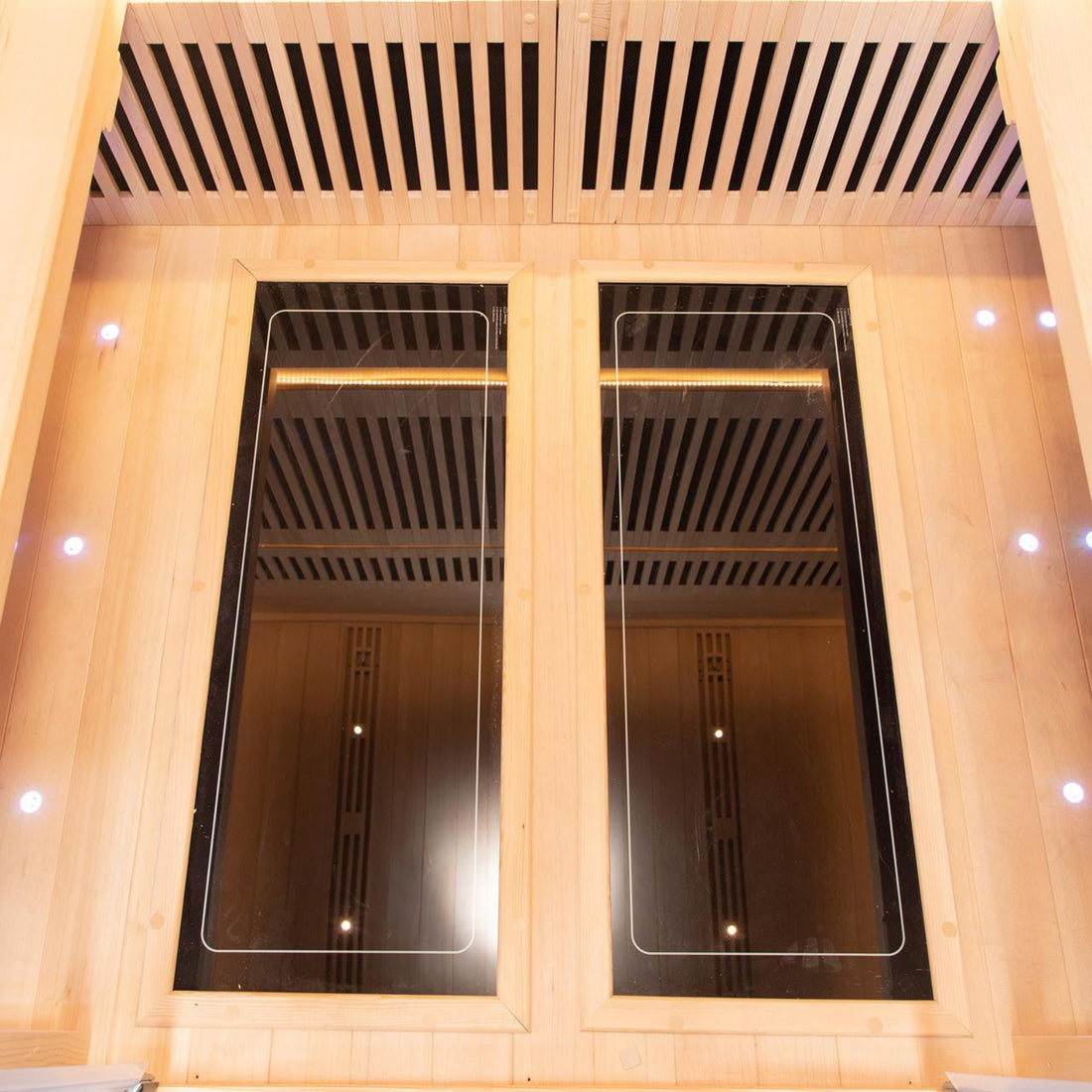 Interior of the Hoito 4-Person Full Spectrum Infrared Sauna with Glass Floor Heaters, featuring two large black infrared panels on a wood-lined wall, small LED spotlights, and ergonomic seating.
