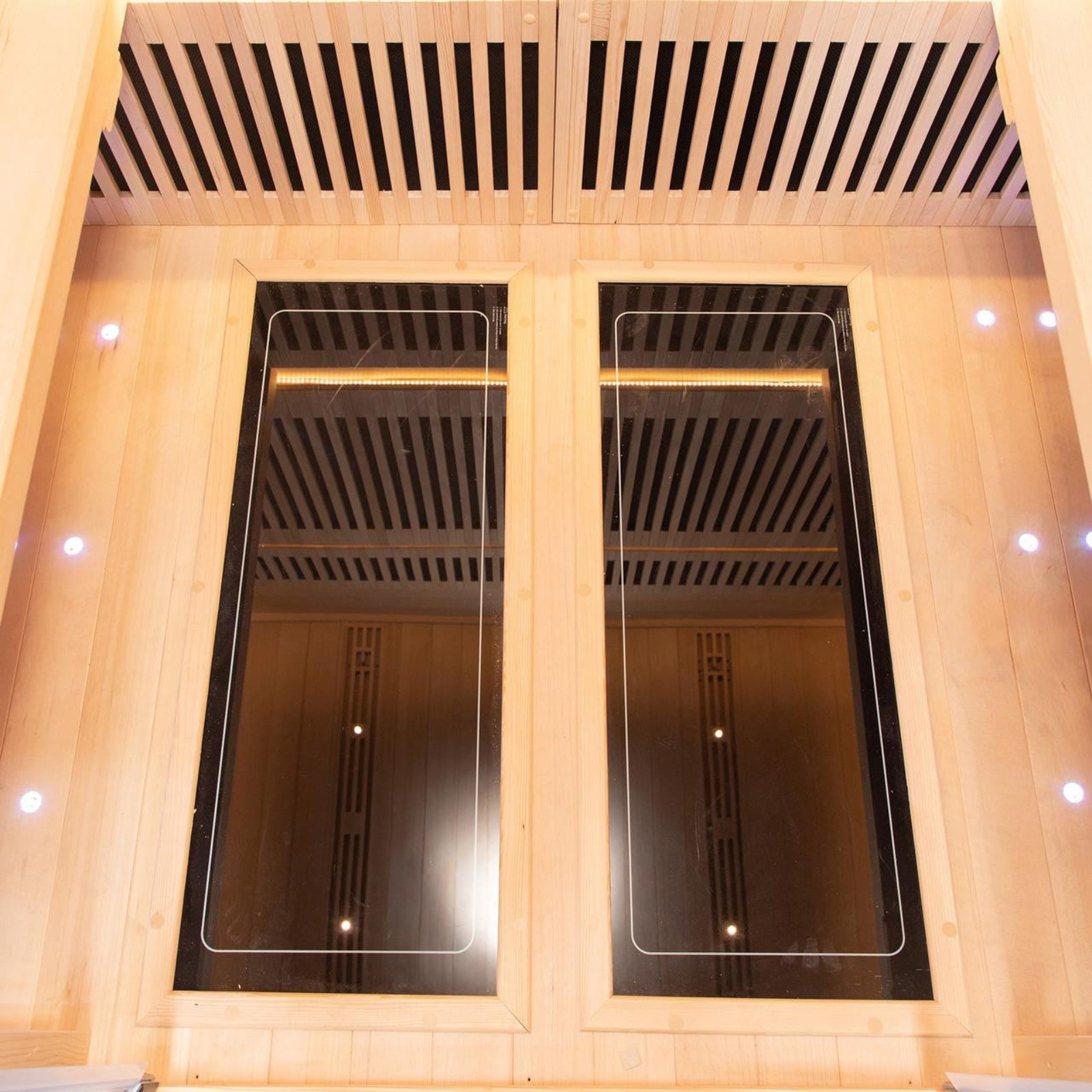 Interior of the Hoito 4-Person Full Spectrum Infrared Sauna with Glass Floor Heaters, featuring two large black infrared panels on a wood-lined wall, small LED spotlights, and ergonomic seating.