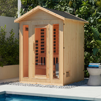 Kiima 3 Person Hybrid Finnish Outdoor Sauna in Natural, a light-colored timber cabin with a glass door, warm-lit interior, wood seating, and lush surroundings by a pool, offering dual heating options.