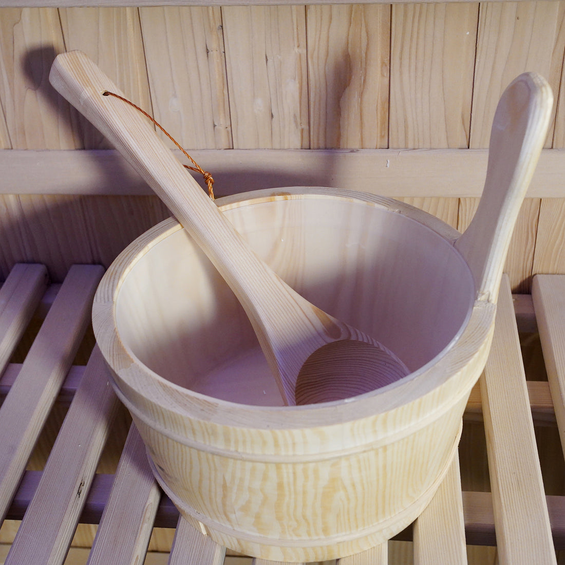 Loyly 3 Person Traditional Finnish Indoor Home Sauna features a pale-wood sauna bucket and ladle on a slatted bench, set against a tongue-and-groove wood wall, enhancing the authentic sauna experience.