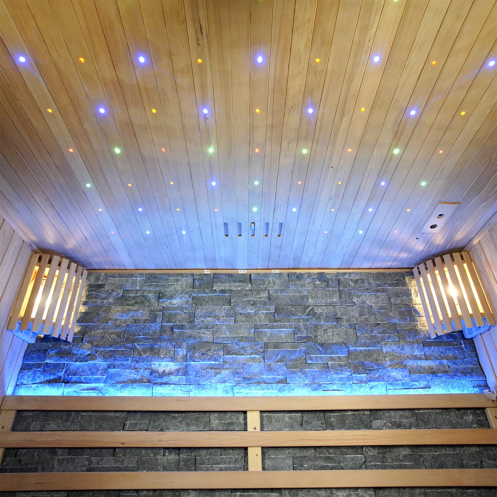 Loyly 3-Person Traditional Finnish Indoor Home Sauna featuring a stone wall with blue LED accents, wooden bench seating, and a star-field effect ceiling, creating a luxurious, calming atmosphere.