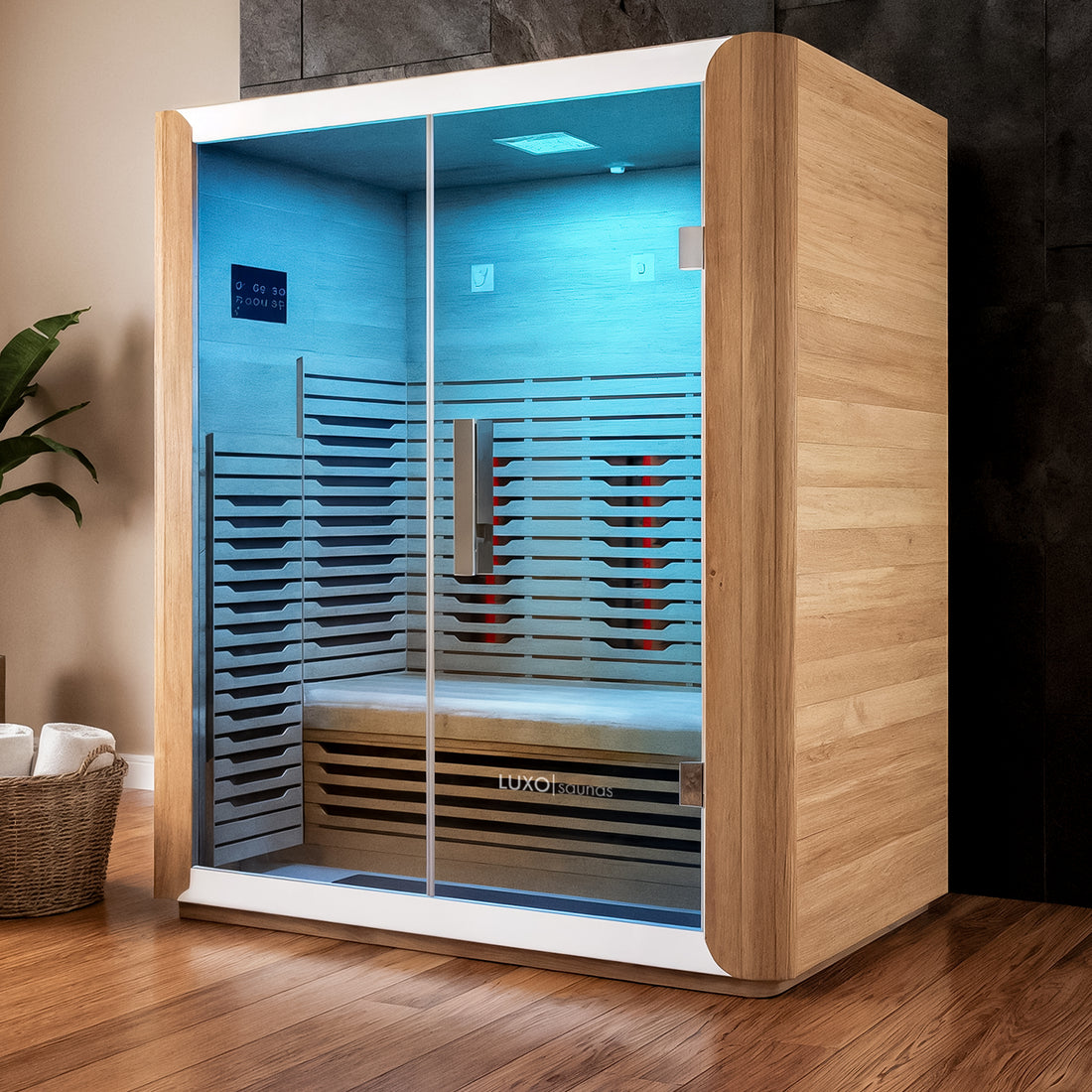 Lue 3 Person Full Spectrum Curved Infrared Indoor Home Sauna, a wooden cabin with glass panels showing a bench inside, softly lit, placed on hardwood floor, beside a towel basket and plant.