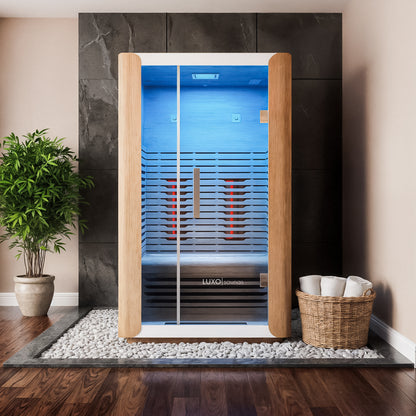 Lue 2 Person Full Spectrum Curved Infrared Indoor Home Sauna with light wood frame, full-height glass door, glowing red panels, set on white pebbles, surrounded by wood and tile flooring.