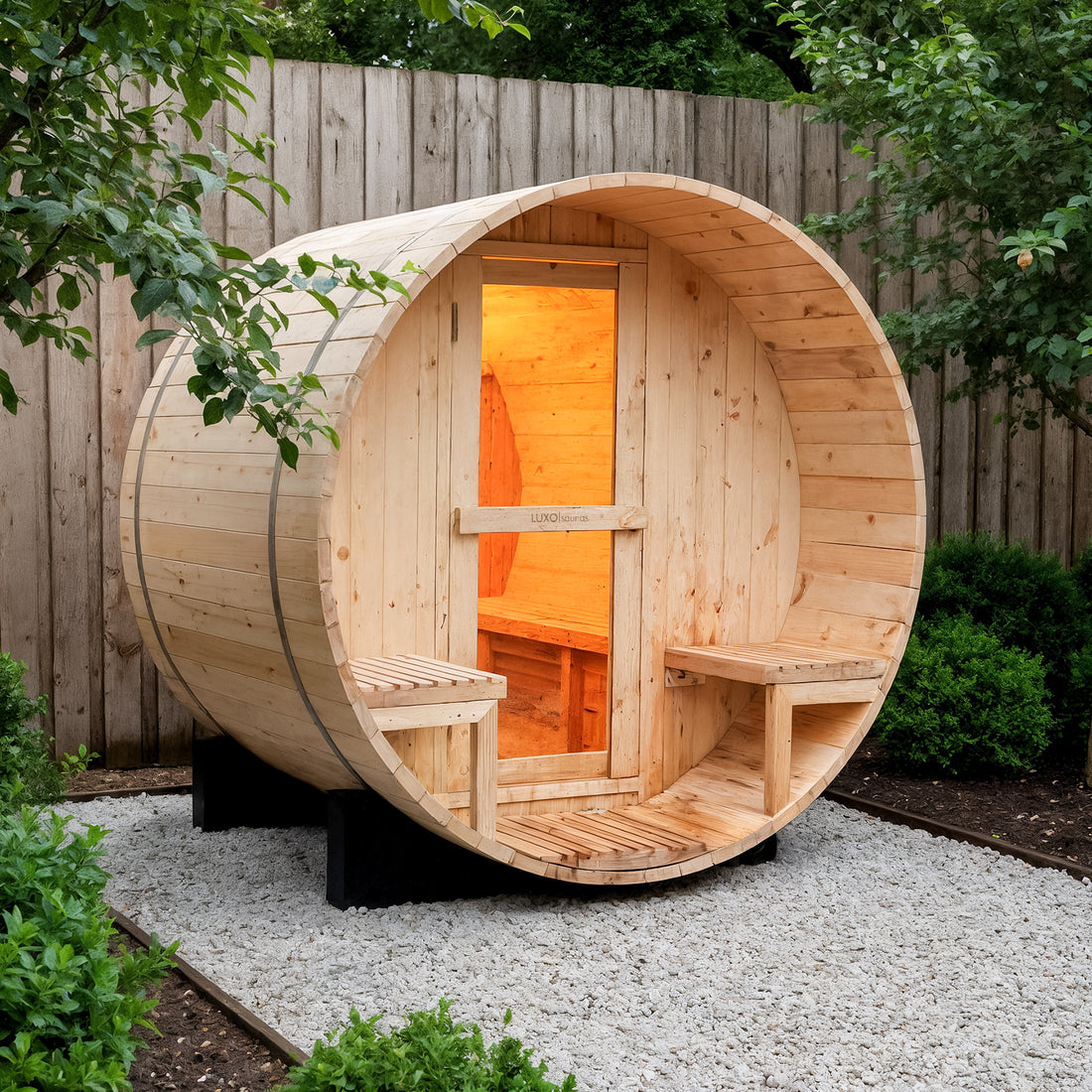 Luola 2 Person Pine Outdoor Barrel Sauna & Harvia Stove in a fenced backyard, showcasing a smooth wooden structure with a glass-paneled door, built-in benches, and warm interior lighting.