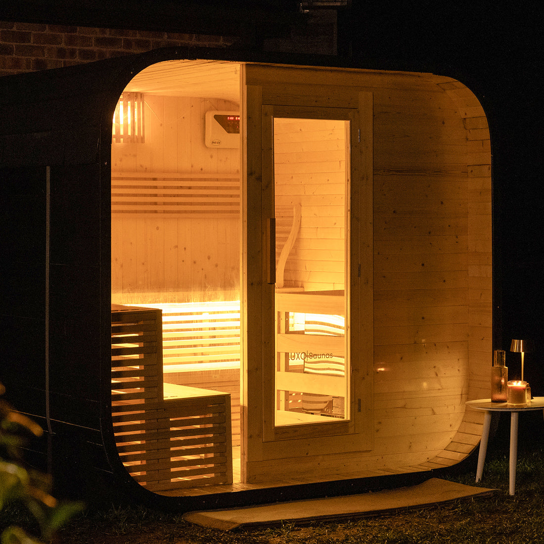 Medika 8 Person Traditional Finnish Outdoor Home Sauna: Barrel-shaped wooden sauna pod glowing warmly at night, featuring interior benches, a digital control unit, and a cozy setup with a side table, candle, glass, and bottle.