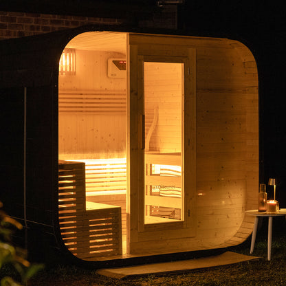 Medika 8 Person Traditional Finnish Outdoor Home Sauna: Barrel-shaped wooden sauna pod glowing warmly at night, featuring interior benches, a digital control unit, and a cozy setup with a side table, candle, glass, and bottle.