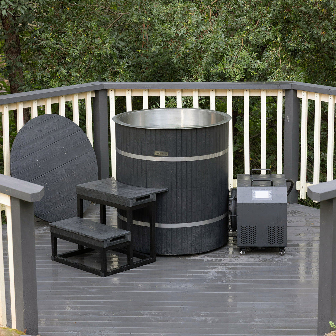 Noni 1-Person Outdoor Ice Bath with 1HP Chiller in Black featuring a round barrel design, stainless-steel interior, dark slatted exterior, matching cover, wooden steps, and a mechanical unit on wheels.