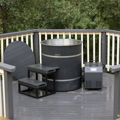 Noni 1-Person Outdoor Ice Bath with 1HP Chiller in Black featuring a round barrel design, stainless-steel interior, dark slatted exterior, matching cover, wooden steps, and a mechanical unit on wheels.