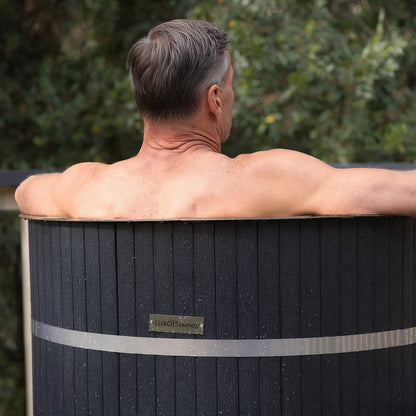 Noni 1-Person Outdoor Ice Bath with 1HP Chiller in Black features a man leaning into a wooden barrel-style tub, showcasing its outdoor setup and premium craftsmanship for cryotherapy and wellness.