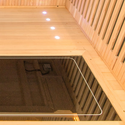 Runko 2 Person Canadian Hemlock Carbon Fibre Infrared Sauna on wooden planks, featuring a sleek design with smooth panels. A blurred hand and face suggest interaction or assembly.