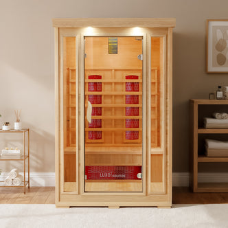 Taavi 2 Person Ceramic Infrared Indoor Home Sauna with pale wood, full-height glass door, visible infrared panels, wooden bench; flanked by a fiddle-leaf fig and snake plant, creating a cozy spa-like ambiance.