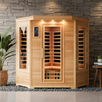 Taavi 4 Person Corner Carbon Fibre Infrared Indoor Sauna with hexagonal shape, glass door, and windows, light wood, recessed lights, and slatted bench, set on pebbles by a stone accent wall.