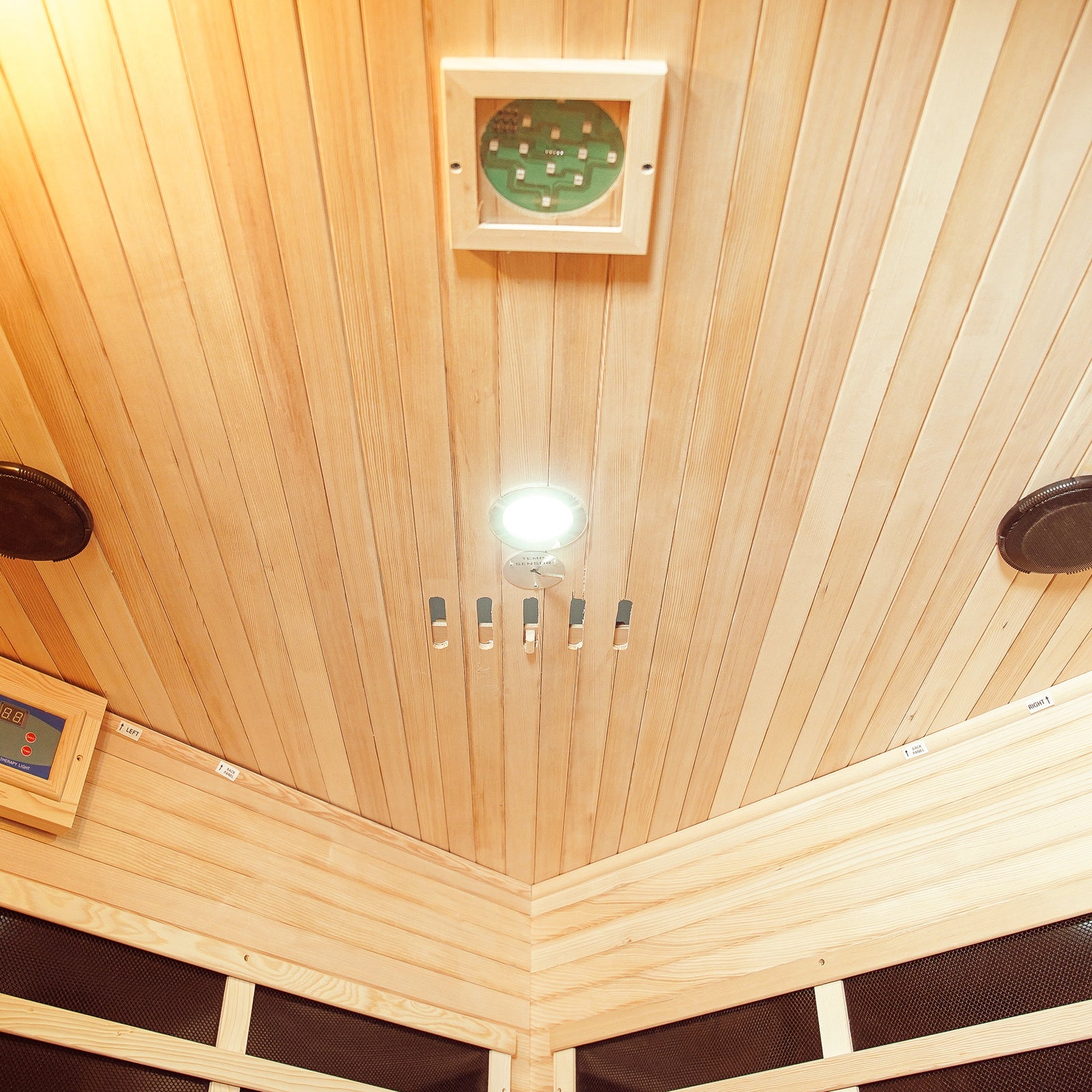Inside view of Taavi 4 Person Corner Carbon Fibre Far Infrared Home Sauna featuring wooden ceiling, recessed light, speakers, thermostat, and digital control panel for a luxurious home spa experience.