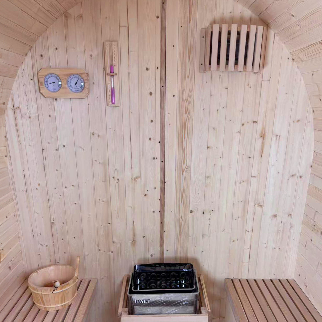 Interior of the Tarni 2-Person Finnish Outdoor Sauna – Fir Wood, featuring pine walls, benches, a sauna bucket, an electric stove with stones, thermometer, hygrometer, sand-timer, and a wooden safety guard.