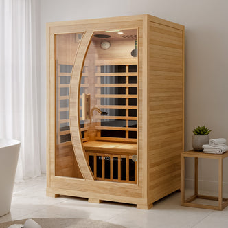 Valo 2 Person Carbon Fibre Indoor Home Sauna with smooth wooden panels, curved glass door, interior bench, and heating panels, set in a serene spa-like bathroom with towels and a potted plant nearby.